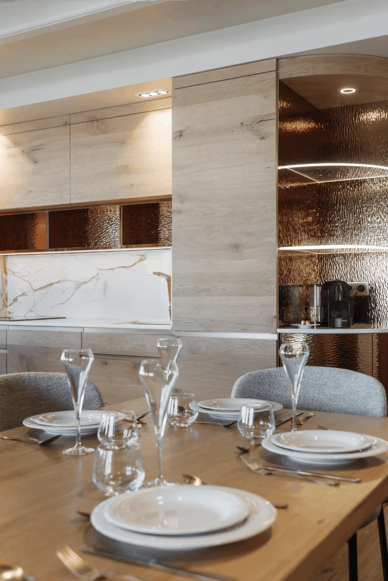 A modern dining room setup with a wooden table set with white plates, clear drinking glasses, and silver cutlery, surrounded by gray upholstered chairs. The background features light wood cabinets and a textured metallic copper/bronze wall panel with built-in lighting.