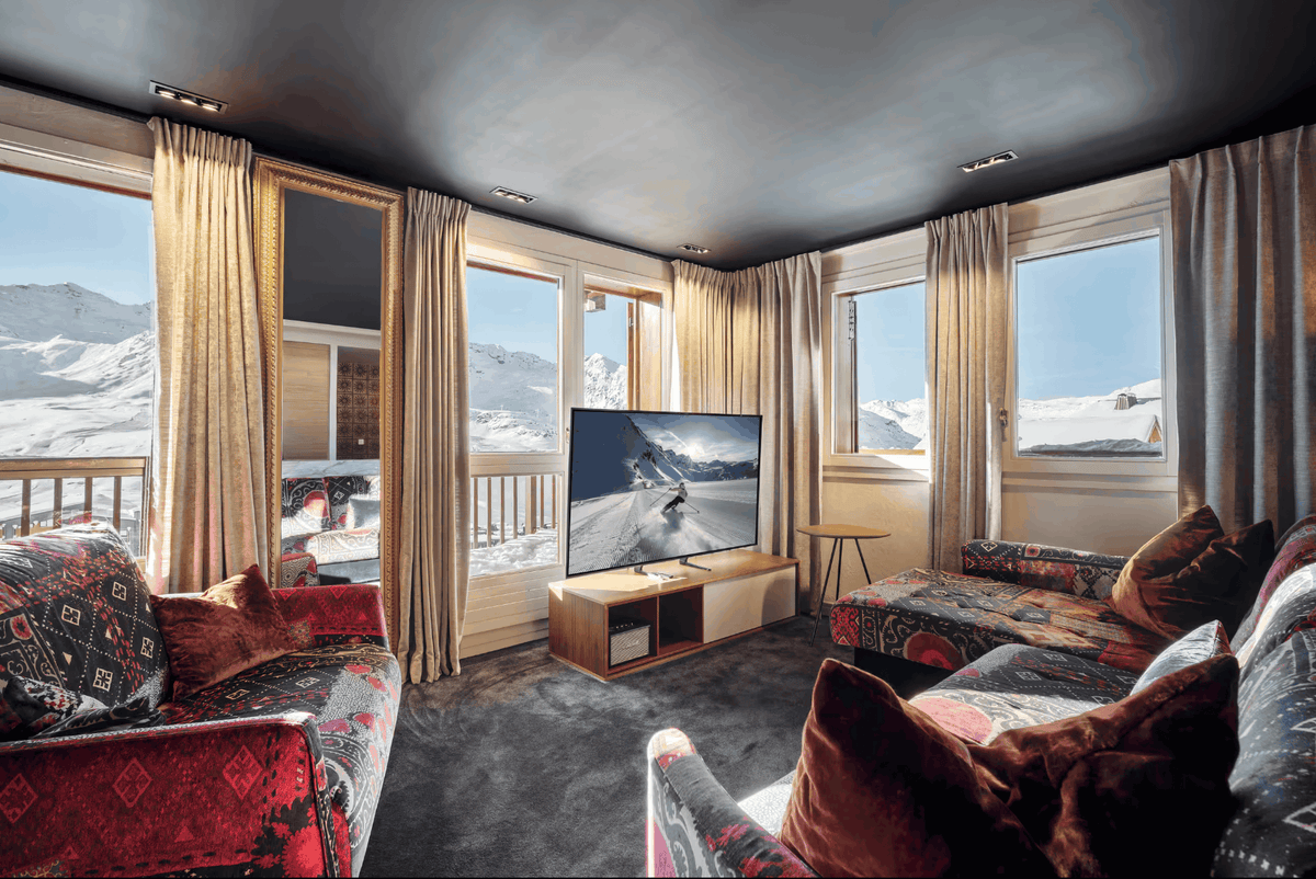 A luxurious ski chalet living room featuring patterned red and black sofas, a wooden media console with a TV, and large windows with gold curtains overlooking a snowy mountain landscape.