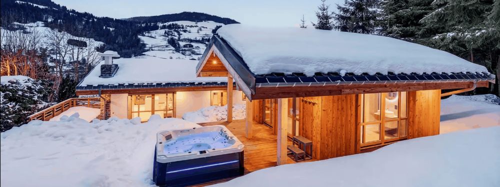A snow-covered chalet with warm glowing windows and a hot tub in the foreground, set in a snowy mountain landscape at dusk.