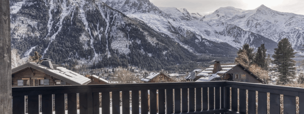A scenic view of snow-covered alpine mountains above a village with wooden chalets, seen from a dark wooden balcony railing in the foreground.