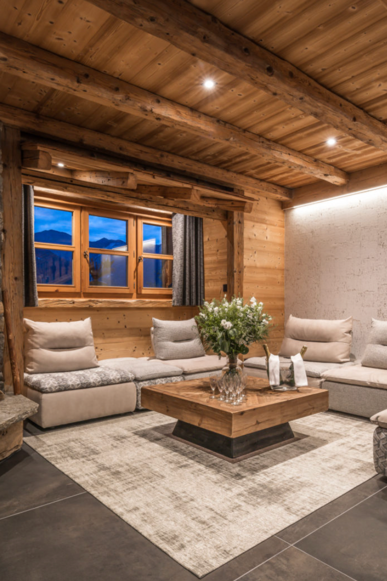 A cozy, rustic mountain chalet living room featuring a U-shaped sectional sofa with beige cushions, a wooden coffee table, a stone fireplace with a roaring fire, and a light-colored textured accent wall.