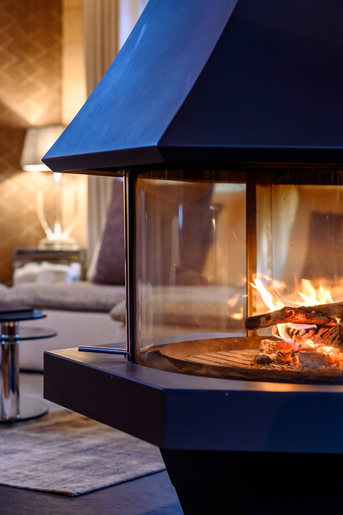 A modern black freestanding fireplace with a glass front and visible flames, set in a warmly lit living room with a sofa and coffee tables in the background.