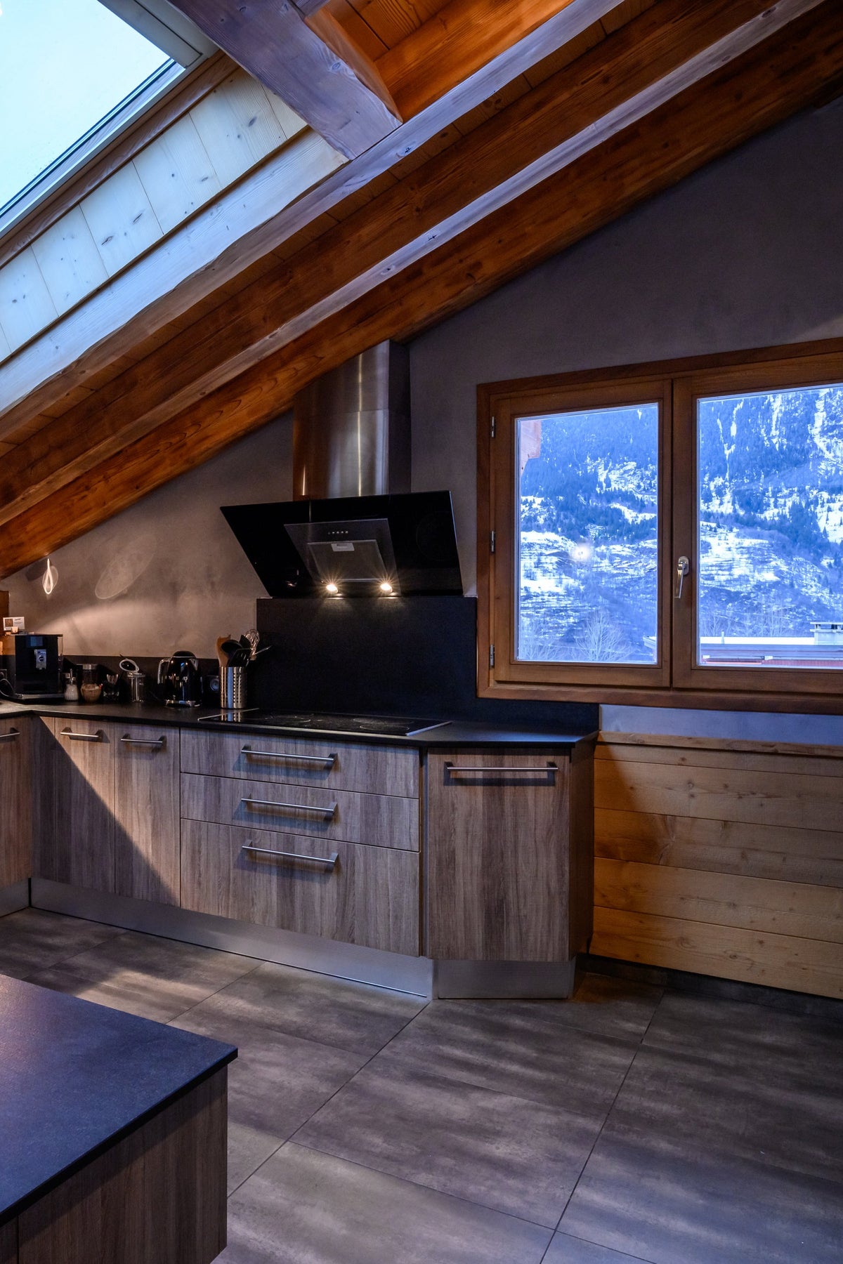 A modern kitchen installed in an attic space with exposed wooden beams and a skylight. The kitchen features light wood cabinetry, dark countertops, a stone-tile backsplash, and a matching island.