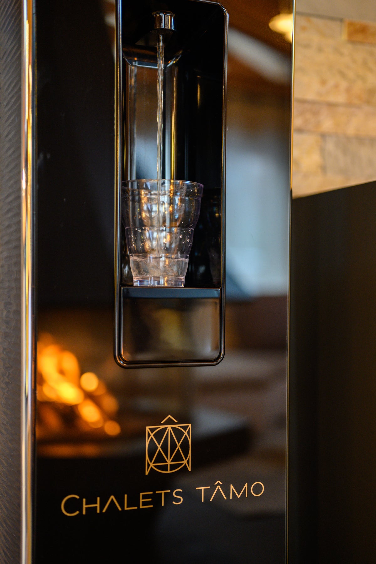A close-up of a sleek black Chalets Tâmo water dispenser with a glass cup under the spout, set in a warm, softly blurred interior environment.