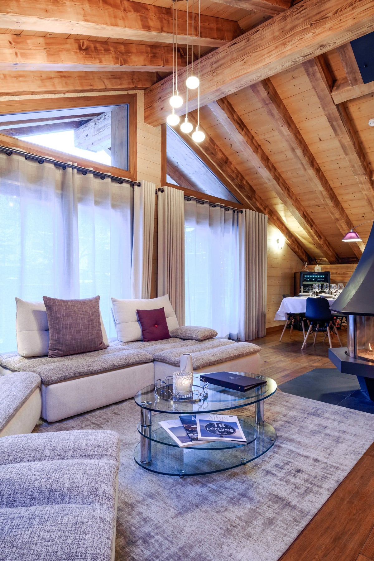 A cozy modern chalet living room featuring a large sectional sofa with purple accent pillows, a glass coffee table, a prominent black wood-burning fireplace with a tall chimney, and a sloped wooden ceiling with exposed beams.