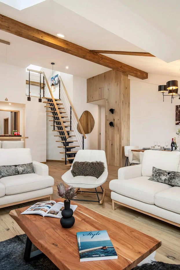 A bright, modern open-plan living room featuring two white sofas with gray patterned pillows, a wooden coffee table, a staircase in the background, and exposed wood ceiling beams.
