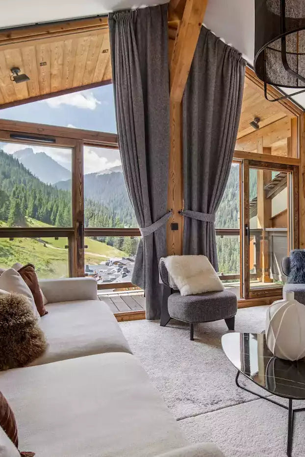 A luxurious ski chalet living room with a large white sectional sofa, dark gray drapes, a glass-top coffee table, and a wall-mounted TV showing a snowy mountain scene. The room features light wood walls, wooden ceiling beams, and large windows overlooking a lush green alpine valley.