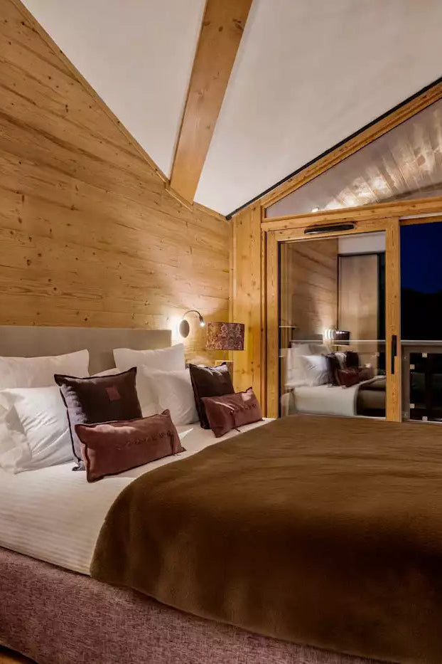 A cozy ski chalet bedroom featuring a large bed with white linens and brown pillows, set against a warm wood-paneled wall. A matching brown blanket covers the bed, and a television mounted on the wall displays a snowy mountain scene.