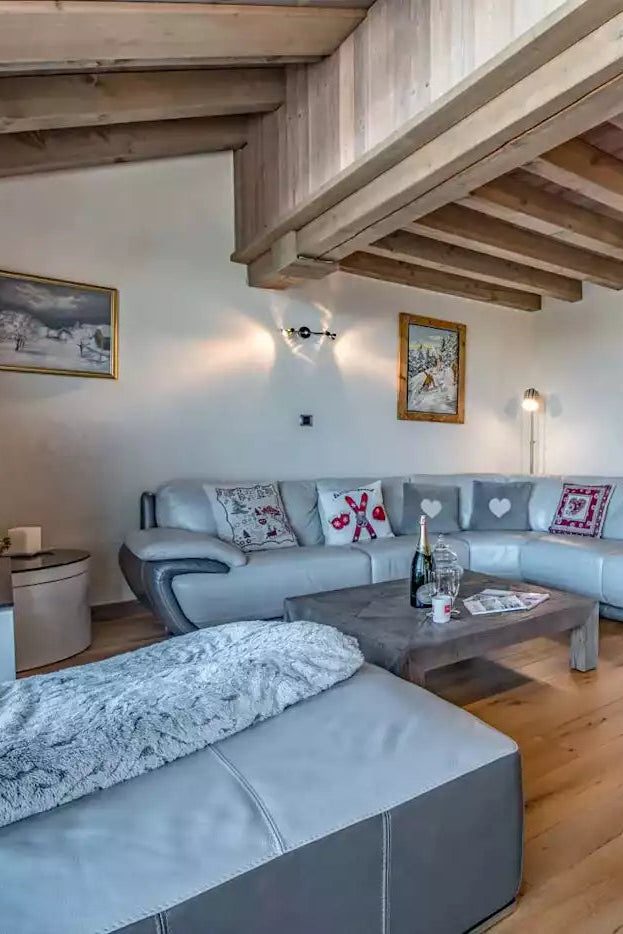 A modern ski chalet living room featuring a light gray sectional sofa, a matching ottoman, a dark wood coffee table, and a large wooden-beamed ceiling. The room has light-colored walls, a gray radiator, and a built-in television stand on the left.
