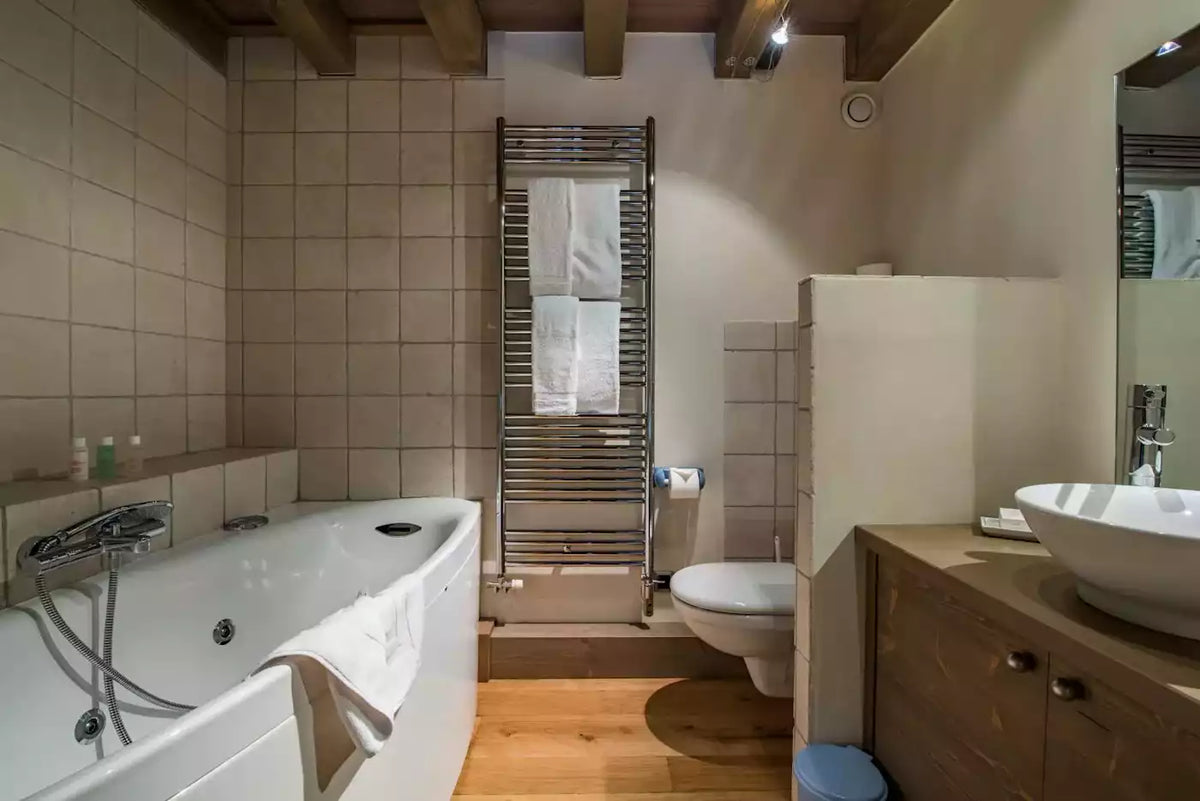 A compact bathroom interior with a white bathtub on the left, a white vessel sink and wooden vanity on the right, a toilet, a towel radiator, and beige tiled walls with exposed wood ceiling beams.
