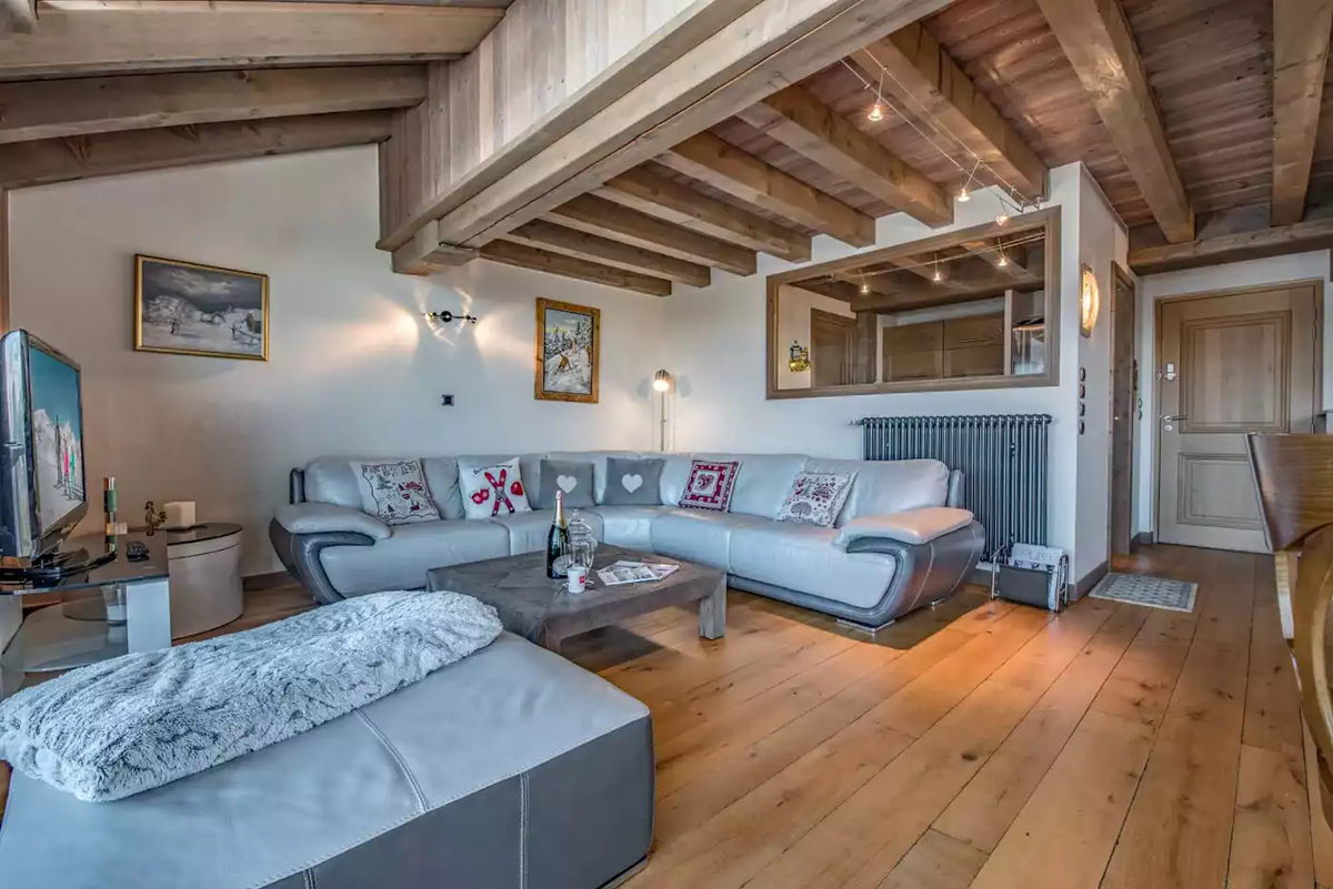 A modern ski chalet living room featuring a light gray sectional sofa, a matching ottoman, a dark wood coffee table, and a large wooden-beamed ceiling. The room has light-colored walls, a gray radiator, and a built-in television stand on the left.