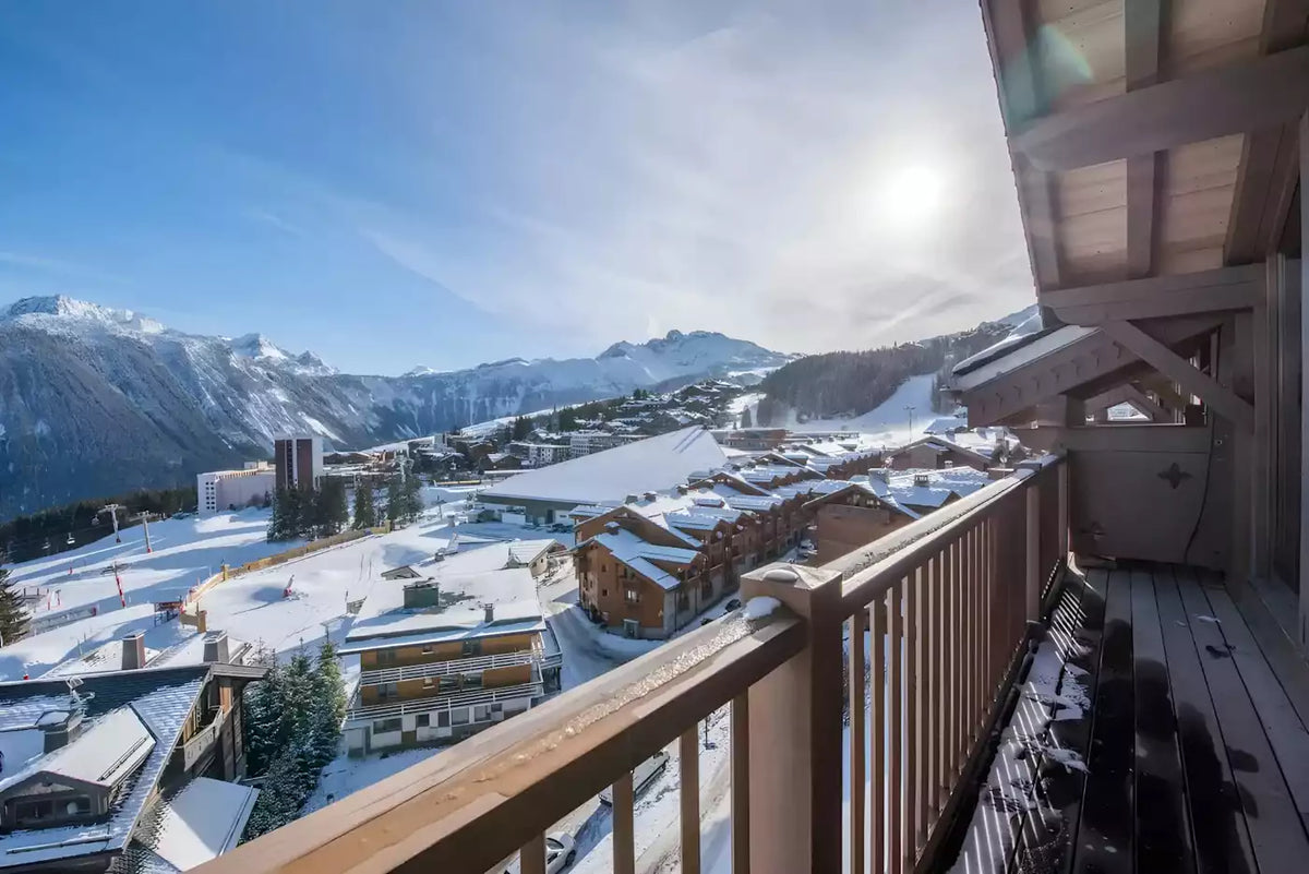 A sunny mountain village with snow-covered roofs and distant peaks, viewed from a wooden balcony with a railing. The bright sun is shining in the sky, and the scene fills the frame.