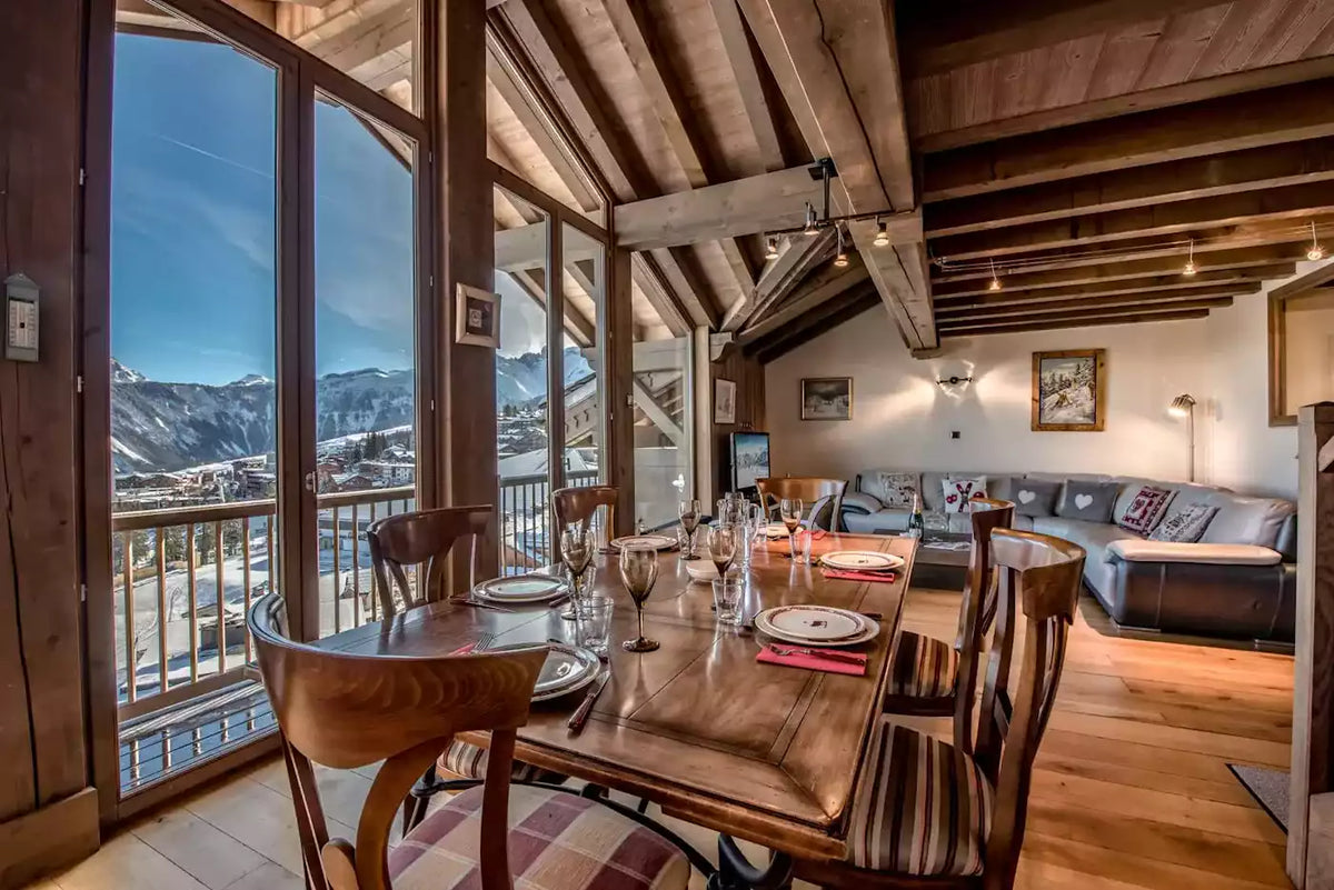 A luxury chalet-style dining room featuring a wooden dining table and chairs set with glassware, overlooking a snowy mountain valley. The background reveals a modern open-plan living room with a gray sectional sofa, wooden ceiling beams, and large floor-to-ceiling windows.