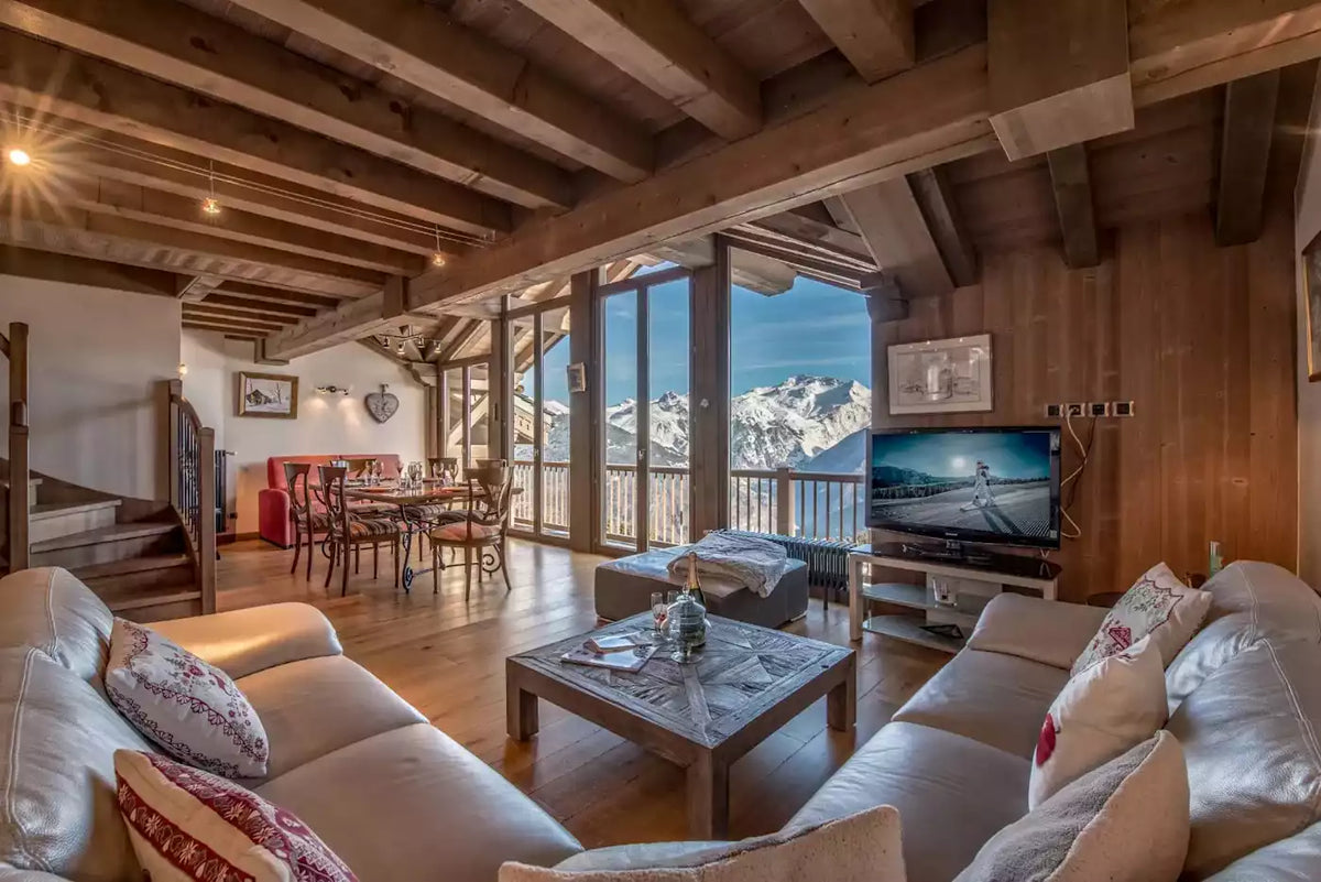 A luxurious chalet-style living room with exposed wooden ceiling beams, cream leather sofas, a marble coffee table, and a large TV displaying a mountain road scene. Large floor-to-ceiling windows offer a stunning view of snow-capped mountains under a blue sky.