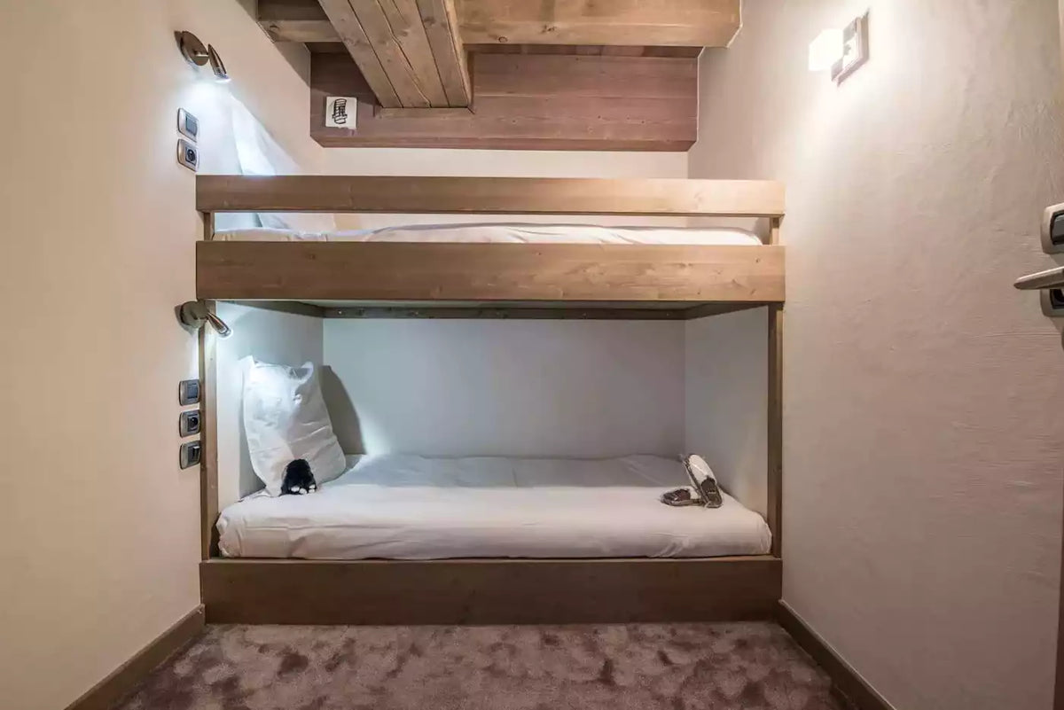 A compact wooden bunk bed with white bedding in a cozy, compact room with warm lighting and textured walls.
