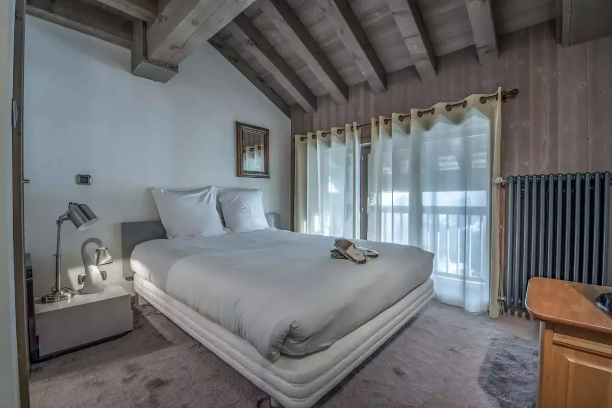 A modern bedroom with a large bed dressed in white bedding, set in a rustic room with exposed wooden ceiling beams, light walls, sheer curtains, and a wooden cabinet in the foreground.