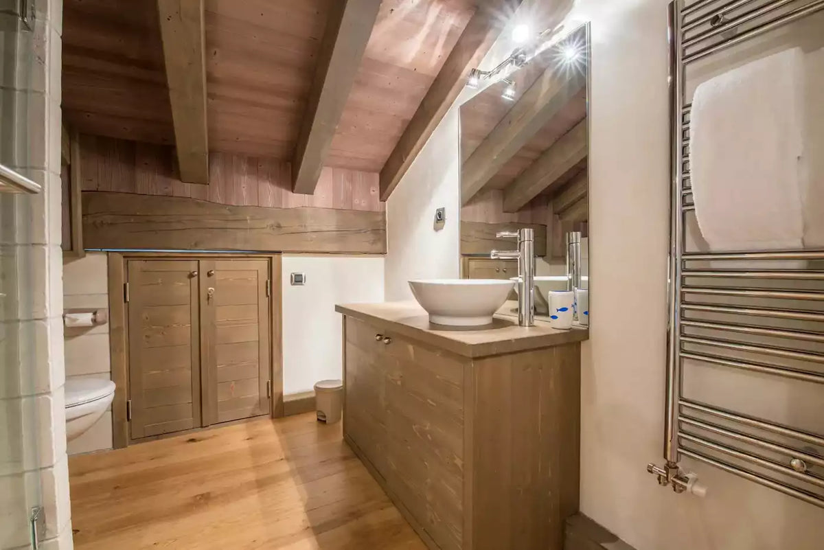 A rustic modern bathroom with light wood ceiling beams, a wooden vanity with a white vessel sink, a large mirror, and a chrome towel radiator on the right.