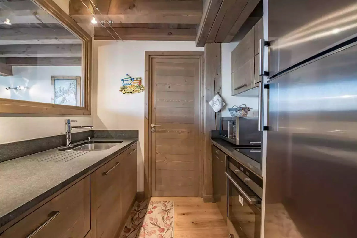A narrow kitchen corridor with wood cabinetry, a stainless steel refrigerator on the right, a sink and countertop on the left, and a wooden door at the end of the passage.