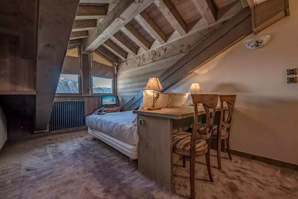 A rustic attic bedroom with exposed wooden beams, a bed with white bedding, a small wooden desk with two chairs, and warm lamps, photographed indoors.