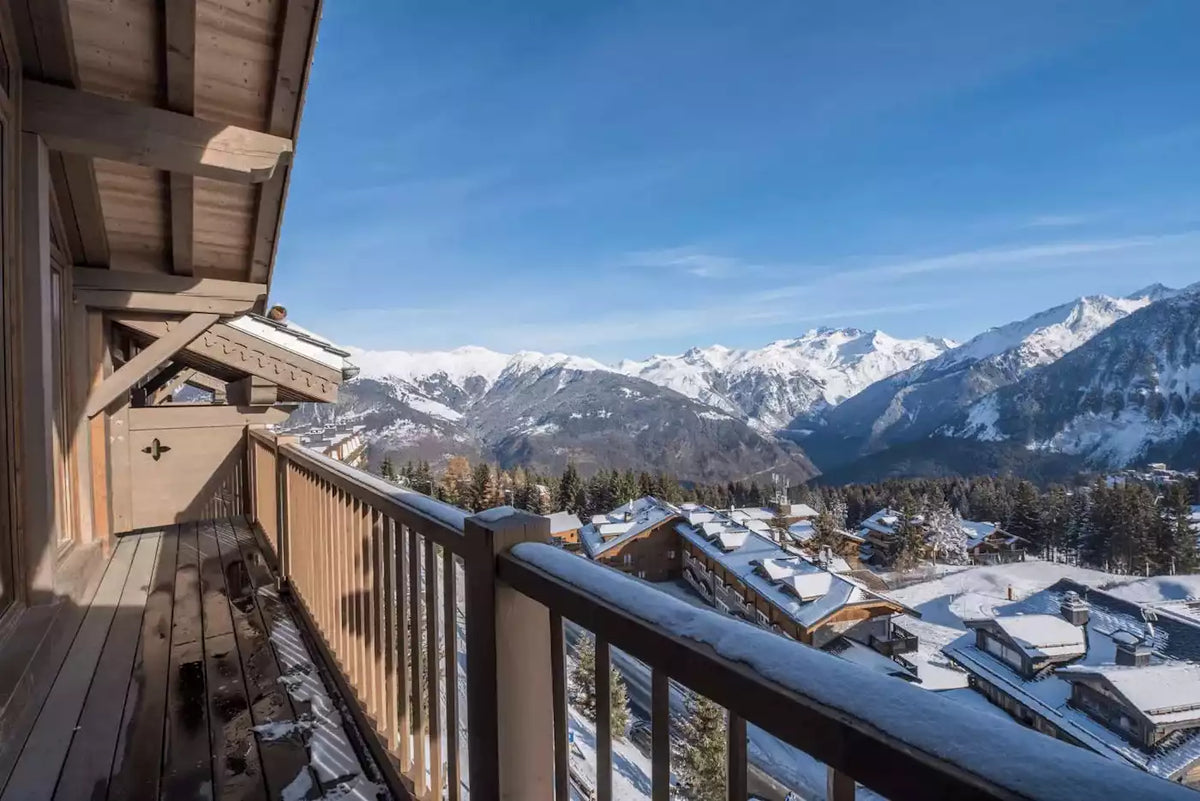A wooden balcony overlooking a snowy mountain village under a clear blue sky, with snow-covered peaks in the background.
