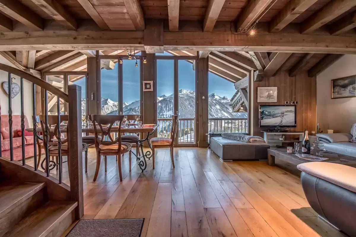 A luxurious alpine chalet interior with wooden floors and ceiling beams, featuring a dining area, living room, and expansive windows showcasing a stunning snow-capped mountain view.