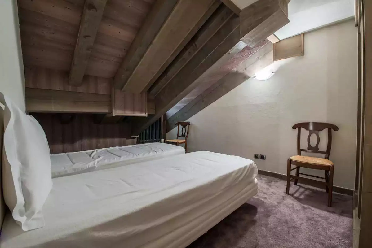 A cozy attic bedroom with two single beds made from a larger bed frame, dressed in white linens. The room features a rustic wooden slatted ceiling, a purple carpet, and a dark wood chair with a woven seat cushion against a light-colored wall.