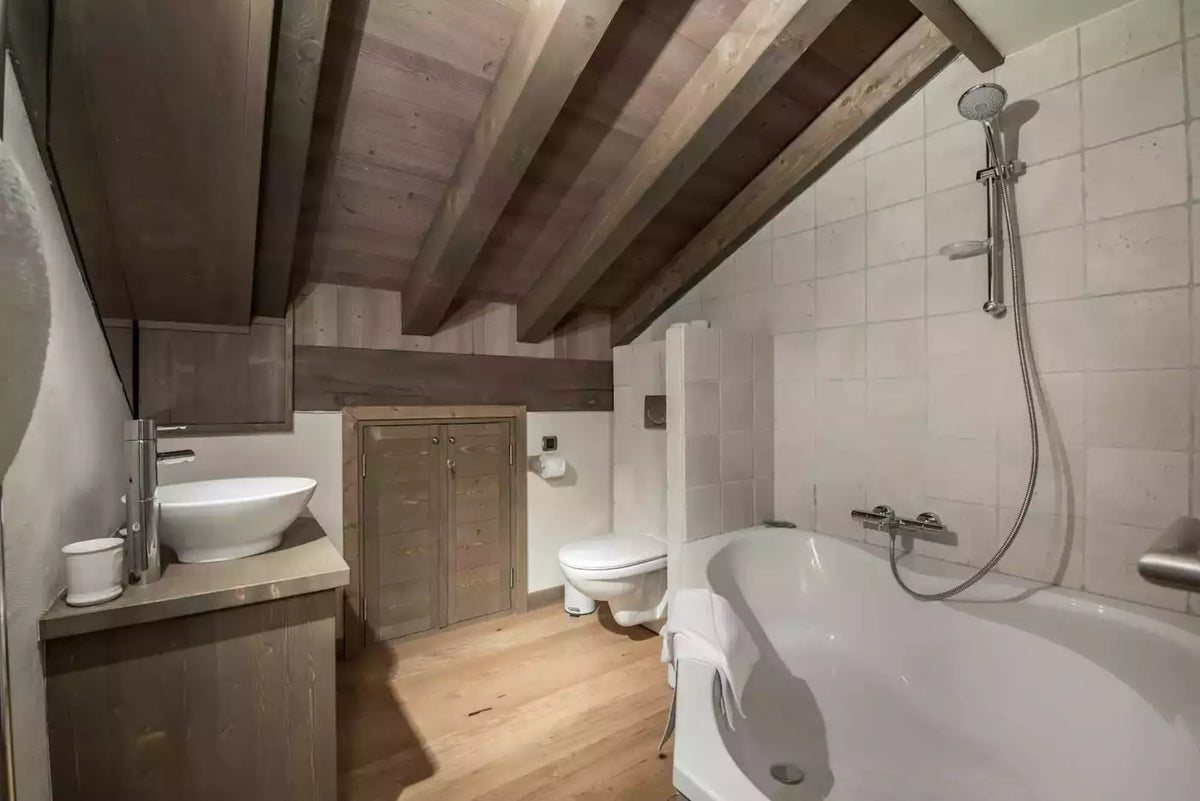 A rustic attic bathroom with exposed wooden ceiling beams, light wood floors, white tiled walls, a white freestanding bathtub with a wall-mounted shower, a white vessel sink on a wood vanity, and a wooden toilet.