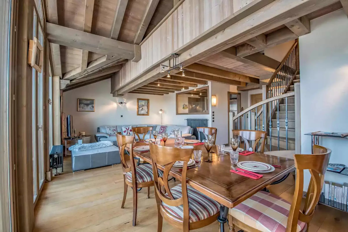 A rustic chalet-style dining room with a wooden dining table and striped upholstered chairs, overlooking a living room with a sofa and a staircase in the background.