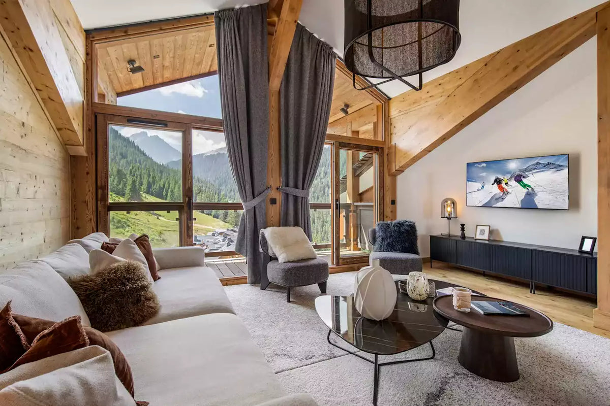 A luxurious ski chalet living room with a large white sectional sofa, dark gray drapes, a glass-top coffee table, and a wall-mounted TV showing a snowy mountain scene. The room features light wood walls, wooden ceiling beams, and large windows overlooking a lush green alpine valley.