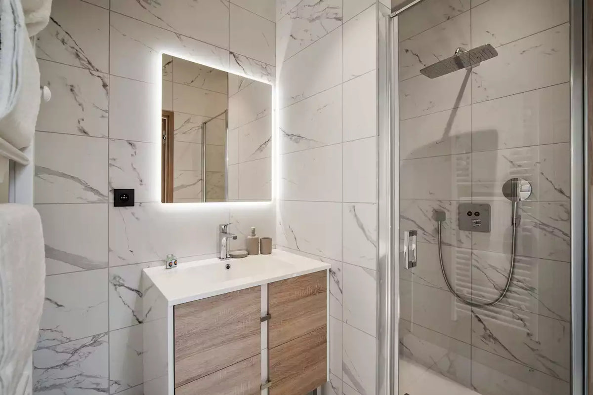 Modern bathroom vanity with an illuminated rectangular mirror, white sink countertop, wood-tone cabinet doors, and a glass shower enclosure with a wall-mounted shower head and handheld shower hose.