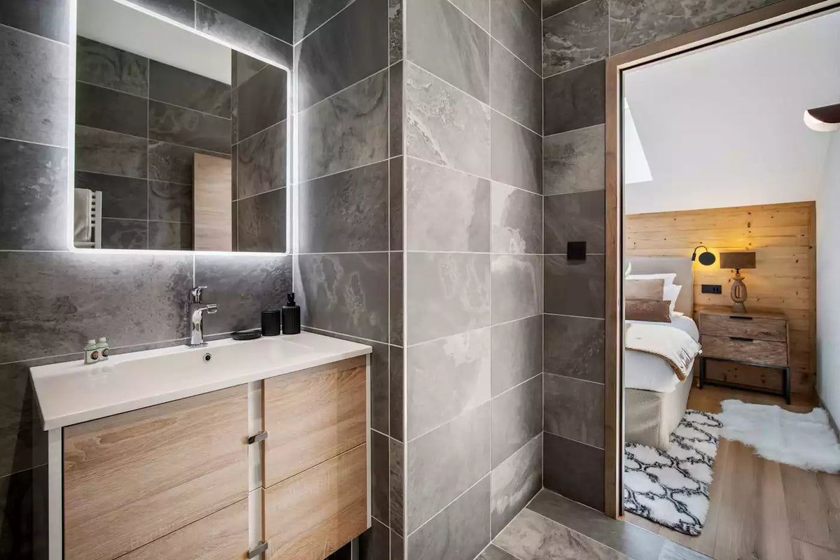 Modern bathroom interior with large gray stone-look tiles, a white countertop and sink cabinet, a backlit rectangular mirror, and an open doorway revealing a bedroom with wood paneling and a bed.