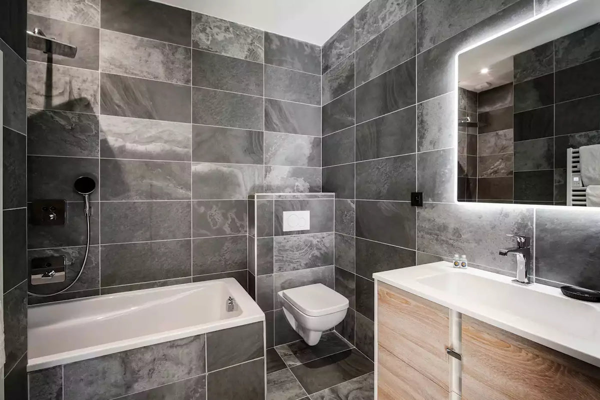 Modern bathroom with dark gray stone-look tiles, a white bathtub, wall-mounted toilet, white sink with light wood vanity, and an illuminated rectangular mirror.