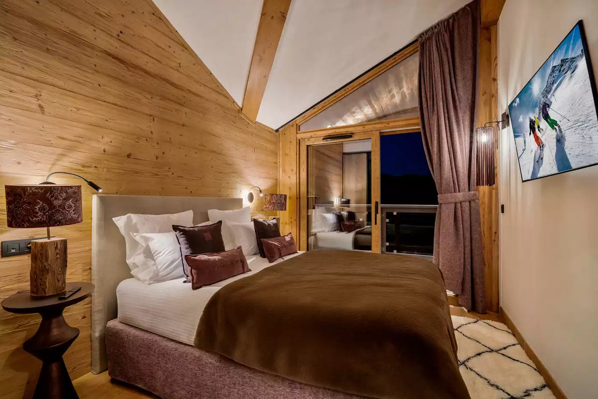 A cozy ski chalet bedroom featuring a large bed with white linens and brown pillows, set against a warm wood-paneled wall. A matching brown blanket covers the bed, and a television mounted on the wall displays a snowy mountain scene.