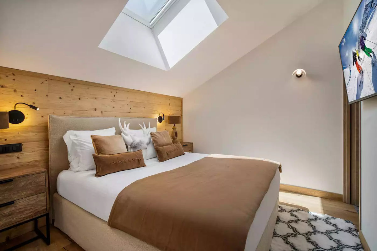 A modern bedroom interior featuring a neatly made bed with white linens and brown pillows, set against a light wood headboard wall. A skylight is visible above the bed, and a wall-mounted TV is shown on the right.