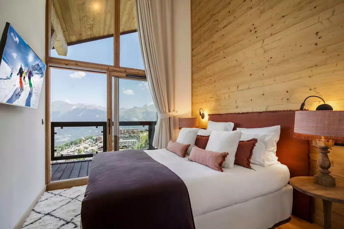 A modern mountain-view bedroom with a large bed dressed in white linens and a dark plum throw, set against a wood-paneled wall. Large windows with beige curtains overlook a scenic lake and mountains, and a wall-mounted TV displays a ski scene.