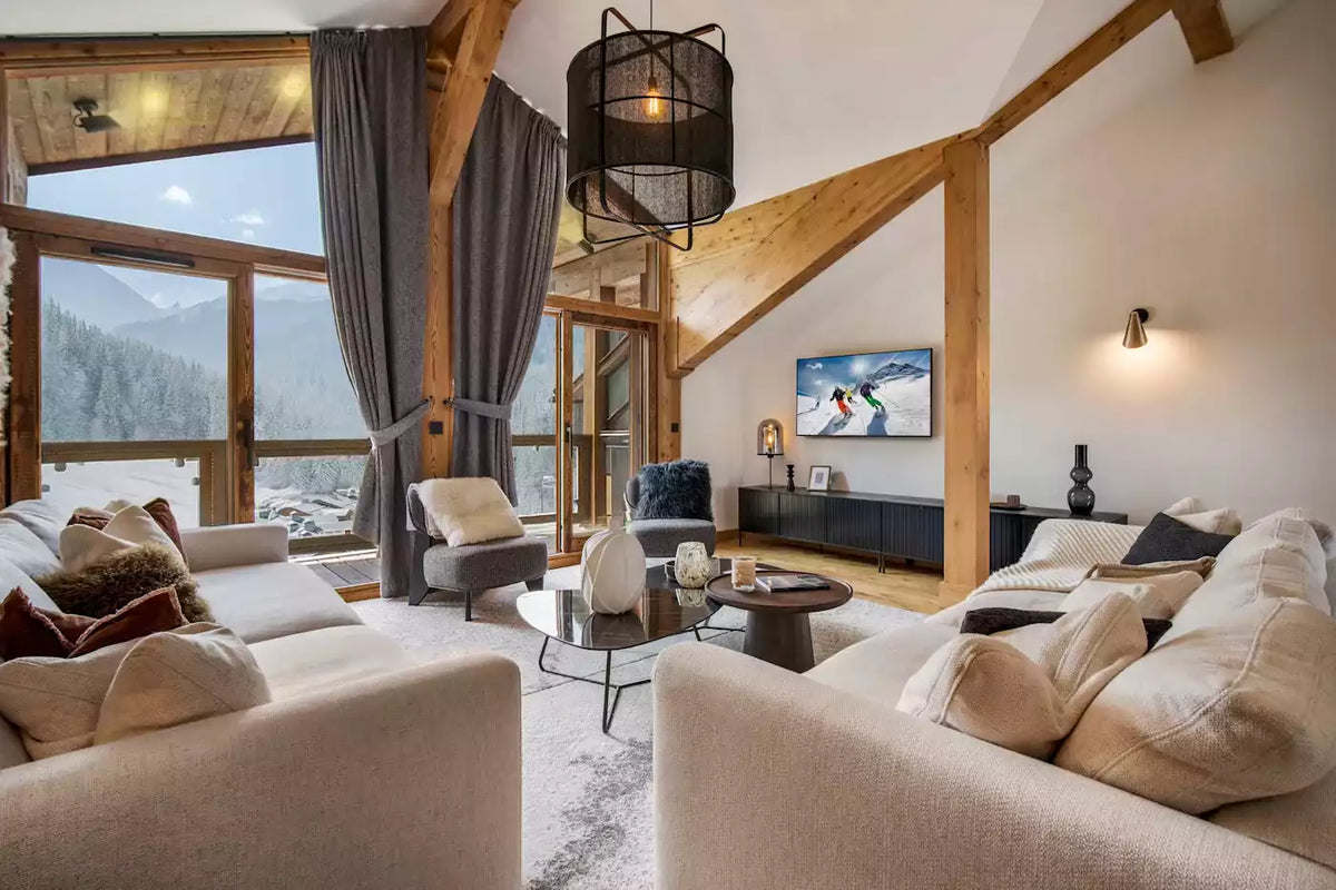 A bright, modern ski chalet living room featuring two cream-colored sofas, a glass coffee table, a wall-mounted TV, and large windows with mountain views.