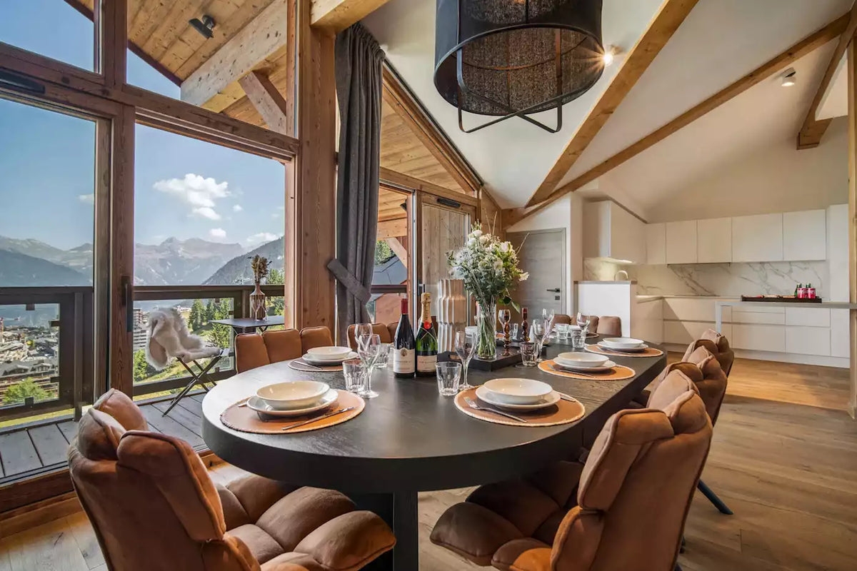 A luxury modern chalet dining room featuring a round dark gray table set with white plates and brown upholstered chairs, overlooking a scenic mountain valley through large floor-to-ceiling windows.