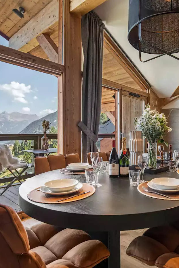 A luxury modern chalet dining room featuring a round dark gray table set with white plates and brown upholstered chairs, overlooking a scenic mountain valley through large floor-to-ceiling windows.