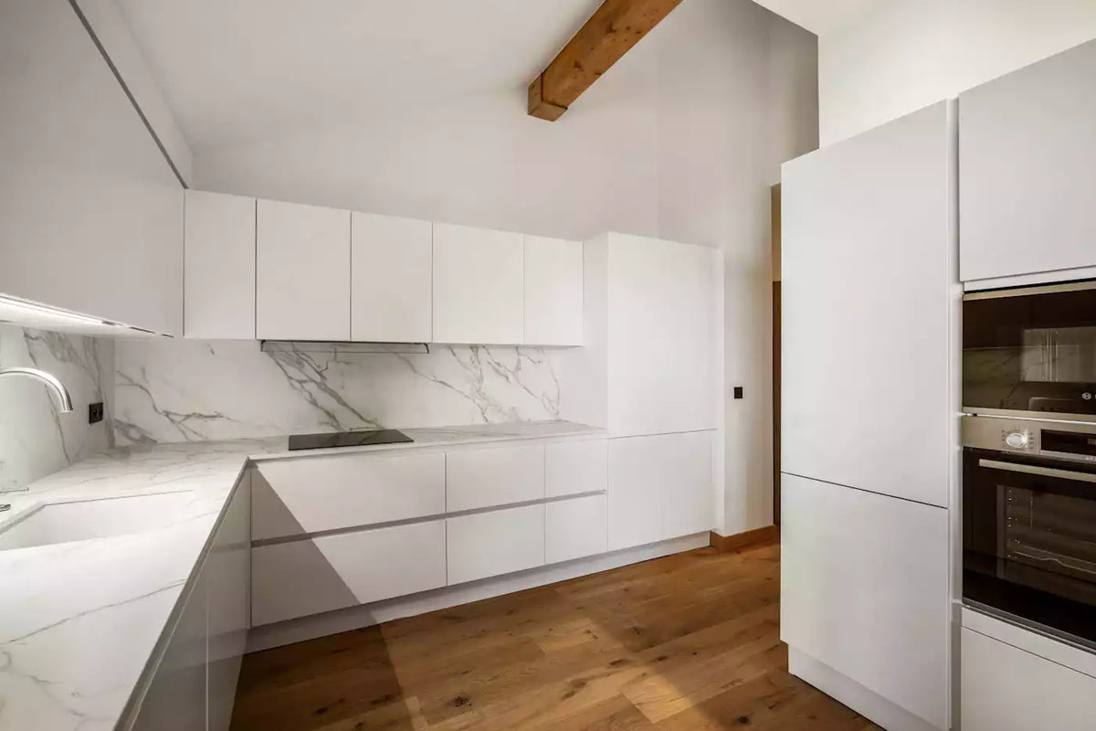 Modern kitchen interior with white flat-panel cabinets, white marble countertops and backsplash, wood flooring, and built-in appliances including a double oven.
