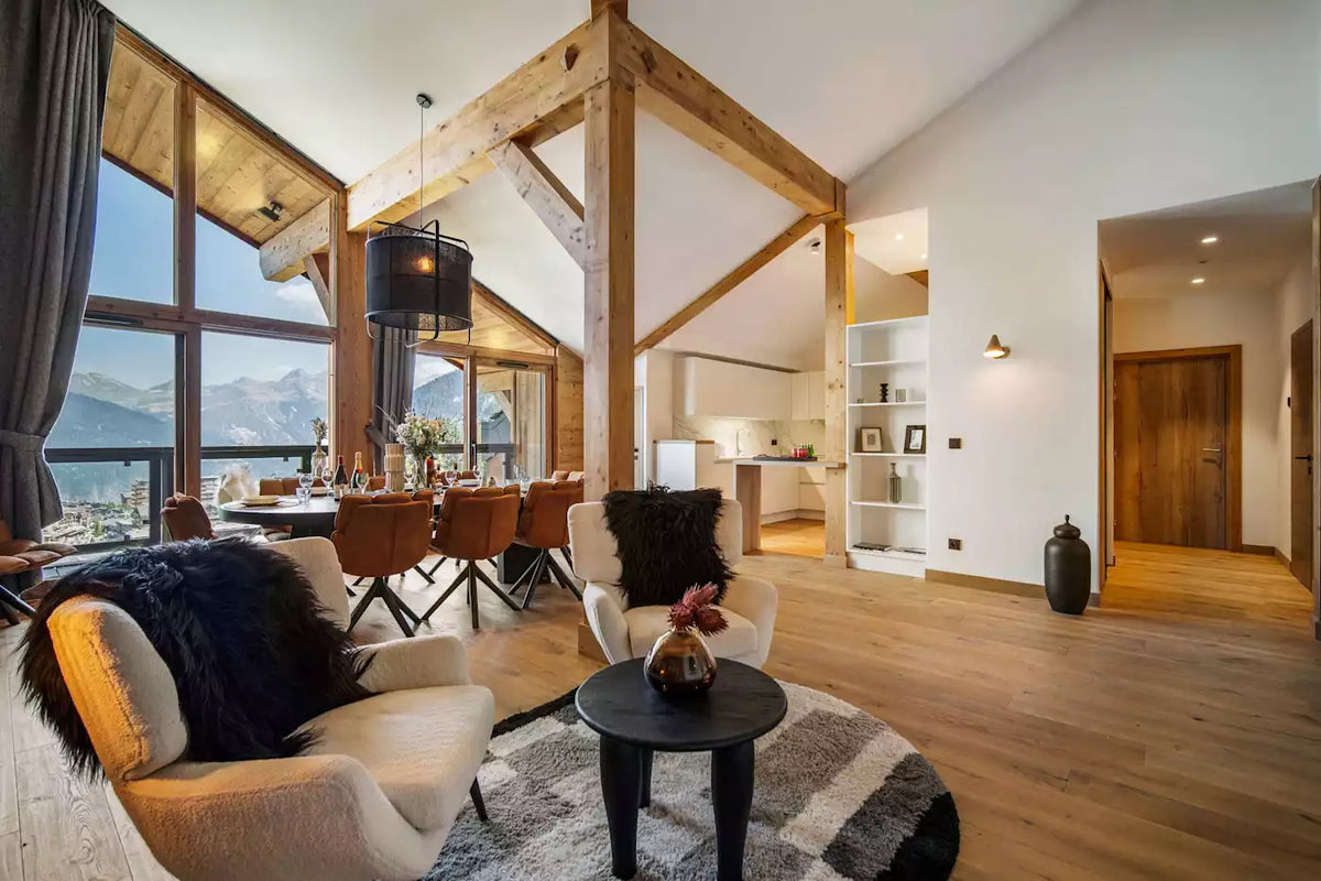 A modern, high-ceilinged chalet living room with exposed wooden beams, large floor-to-ceiling windows, and a mountain view. The room features a cream-colored lounge chair with a black fur throw, a matching armchair, a round black coffee table, and a gray patterned rug.