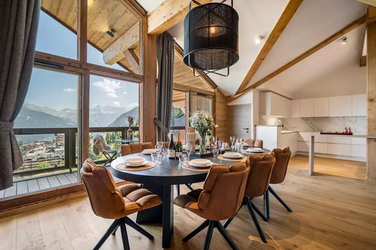 A luxury modern chalet interior featuring a round dark dining table surrounded by brown leather chairs, set under a black pendant light. Large windows offer a stunning view of mountains and a village, while a white kitchen is visible in the background.