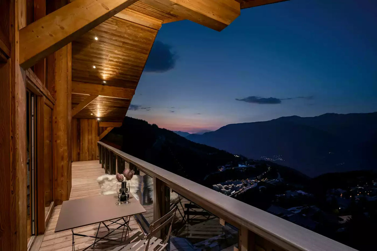 A modern wooden chalet balcony at dusk overlooking a mountain valley and ski village, with a table and chairs on the deck and a deep blue evening sky above.