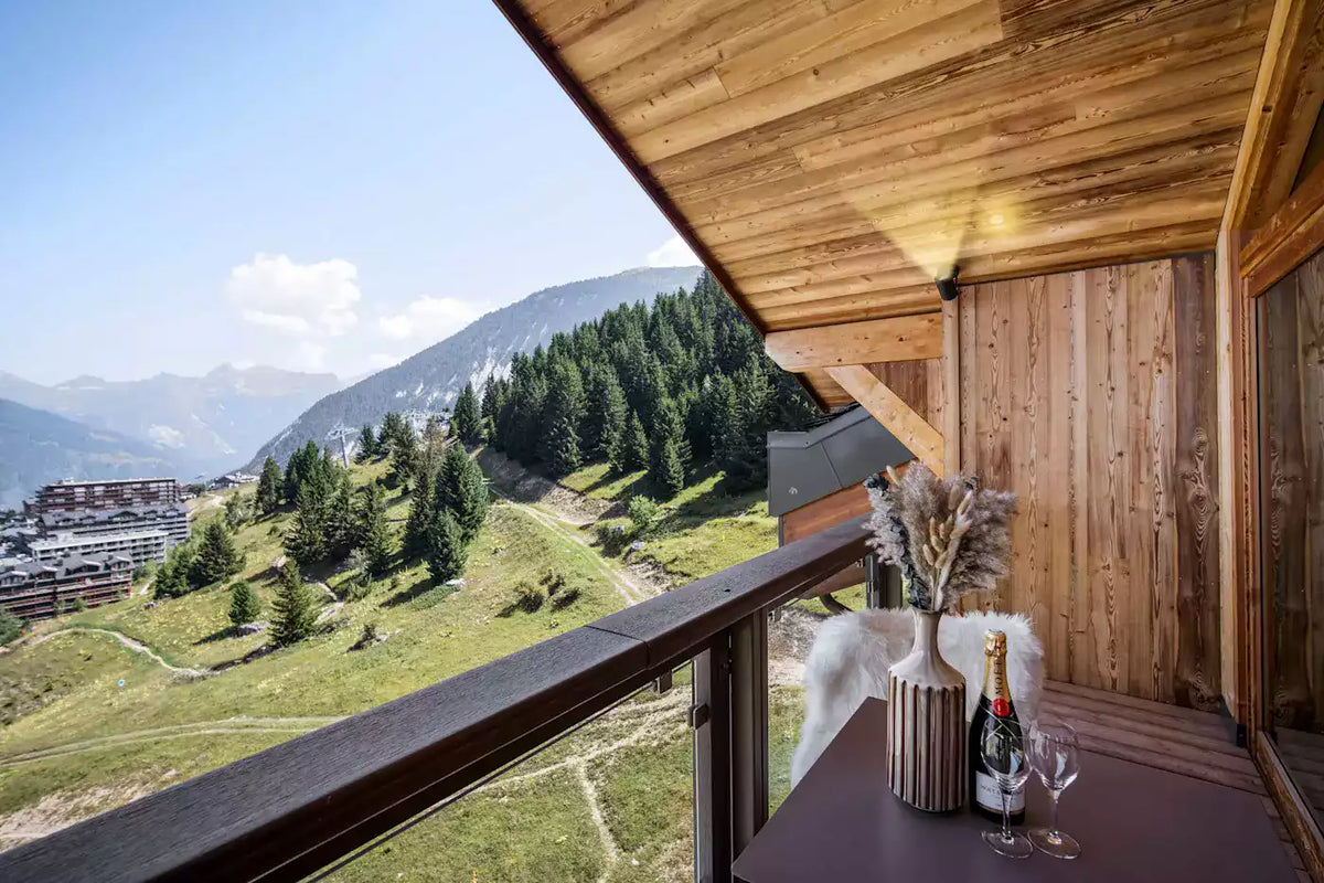 A luxury wooden balcony overlooking a lush green mountain valley with pine trees and distant ski resort buildings under a clear blue sky. A small table on the balcony holds a champagne bottle, a glass, and decorative dried grass in a vase.