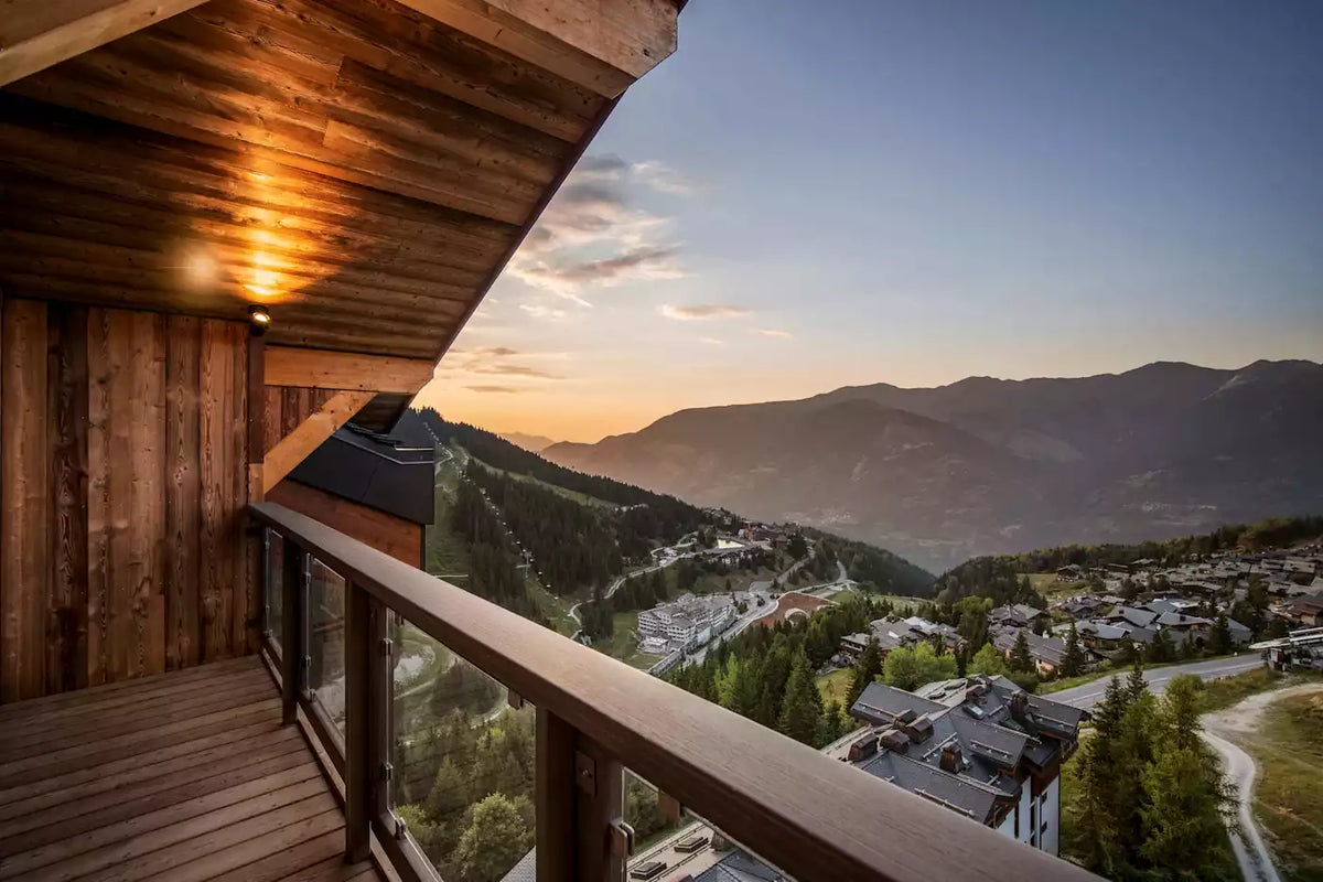 A wooden balcony overlooking a mountain village at sunset, with pine-covered slopes and distant peaks under a blue sky.