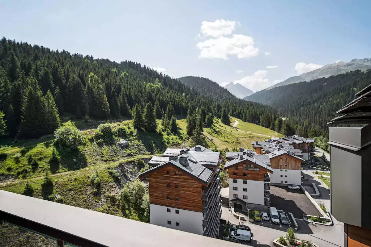 Alpine mountain village scene with chalets, a forested hillside, and distant peaks under a blue sky. A balcony railing is visible in the foreground.