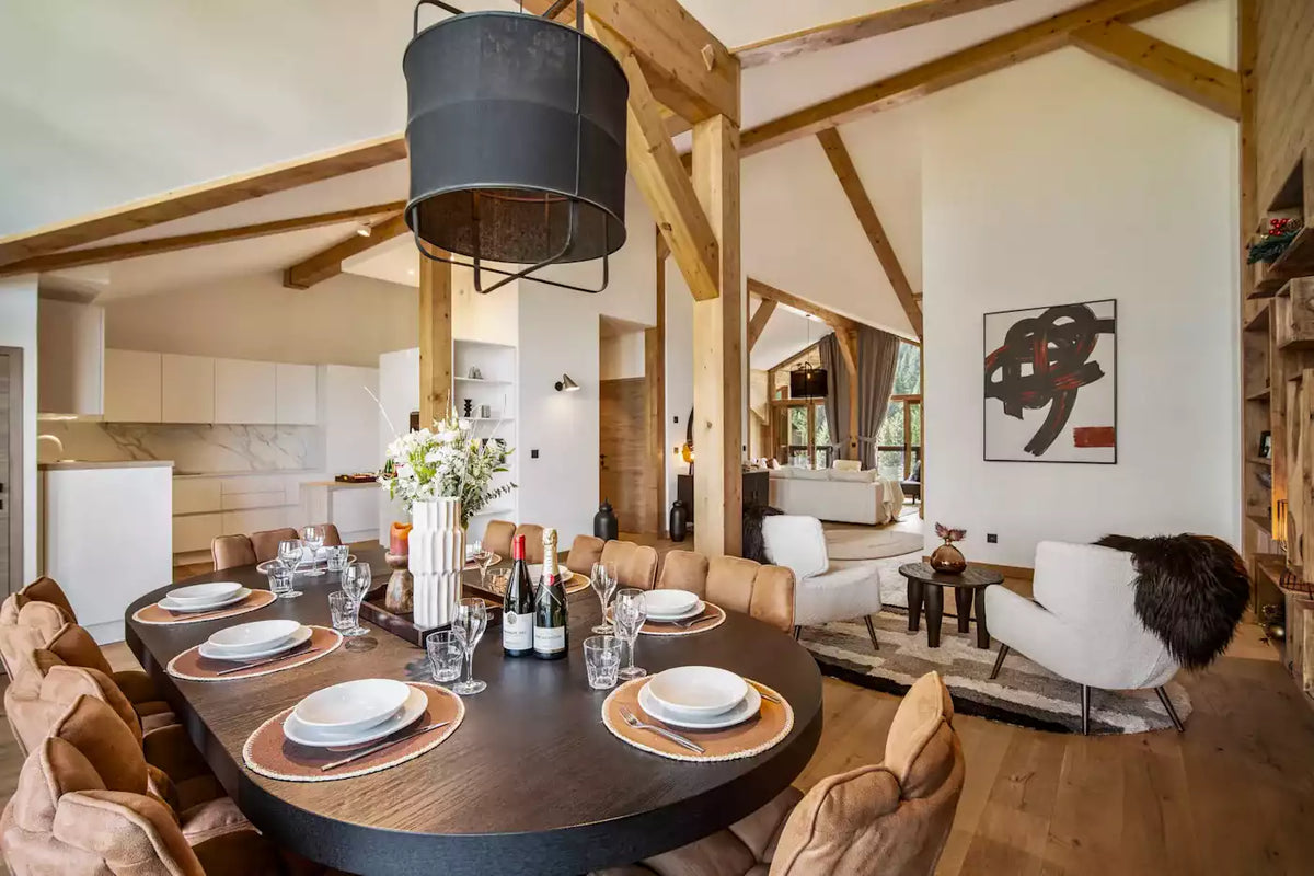 A modern dining room with a dark oval table set for six, surrounded by brown upholstered chairs. The room features exposed wooden beams, a large black pendant light, and an open-plan layout leading to a kitchen and living area.