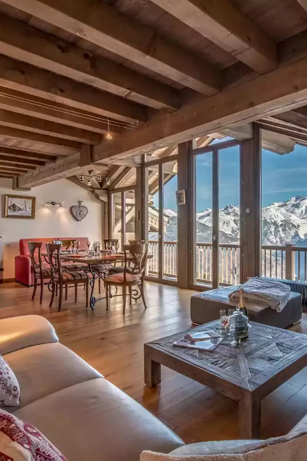A luxurious chalet-style living room with exposed wooden ceiling beams, cream leather sofas, a marble coffee table, and a large TV displaying a mountain road. Large floor-to-ceiling windows offer a stunning view of snow-capped mountains under a blue sky.