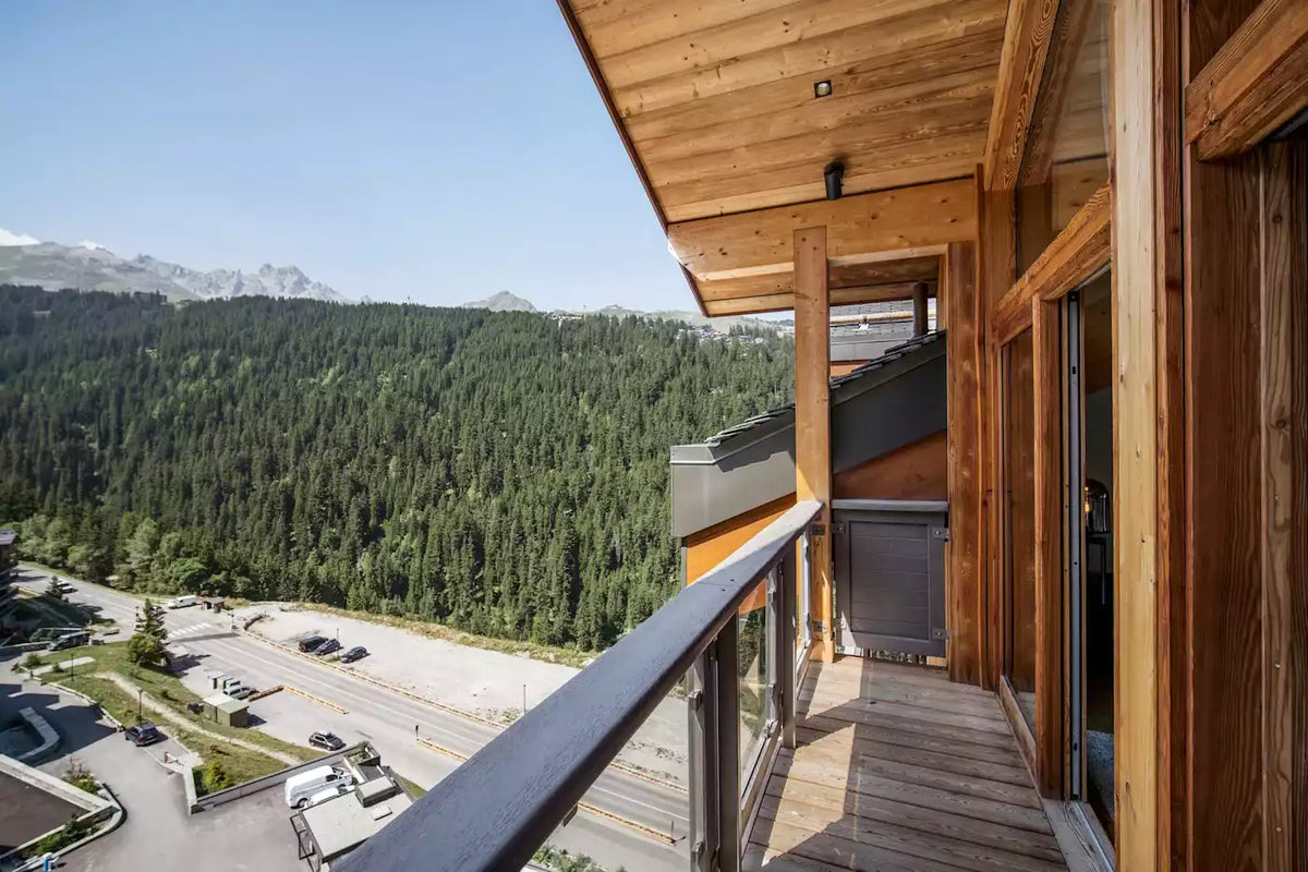 A modern wooden balcony with a glass railing overlooking a vast green mountain forest and distant snow-capped peaks under a clear blue sky.