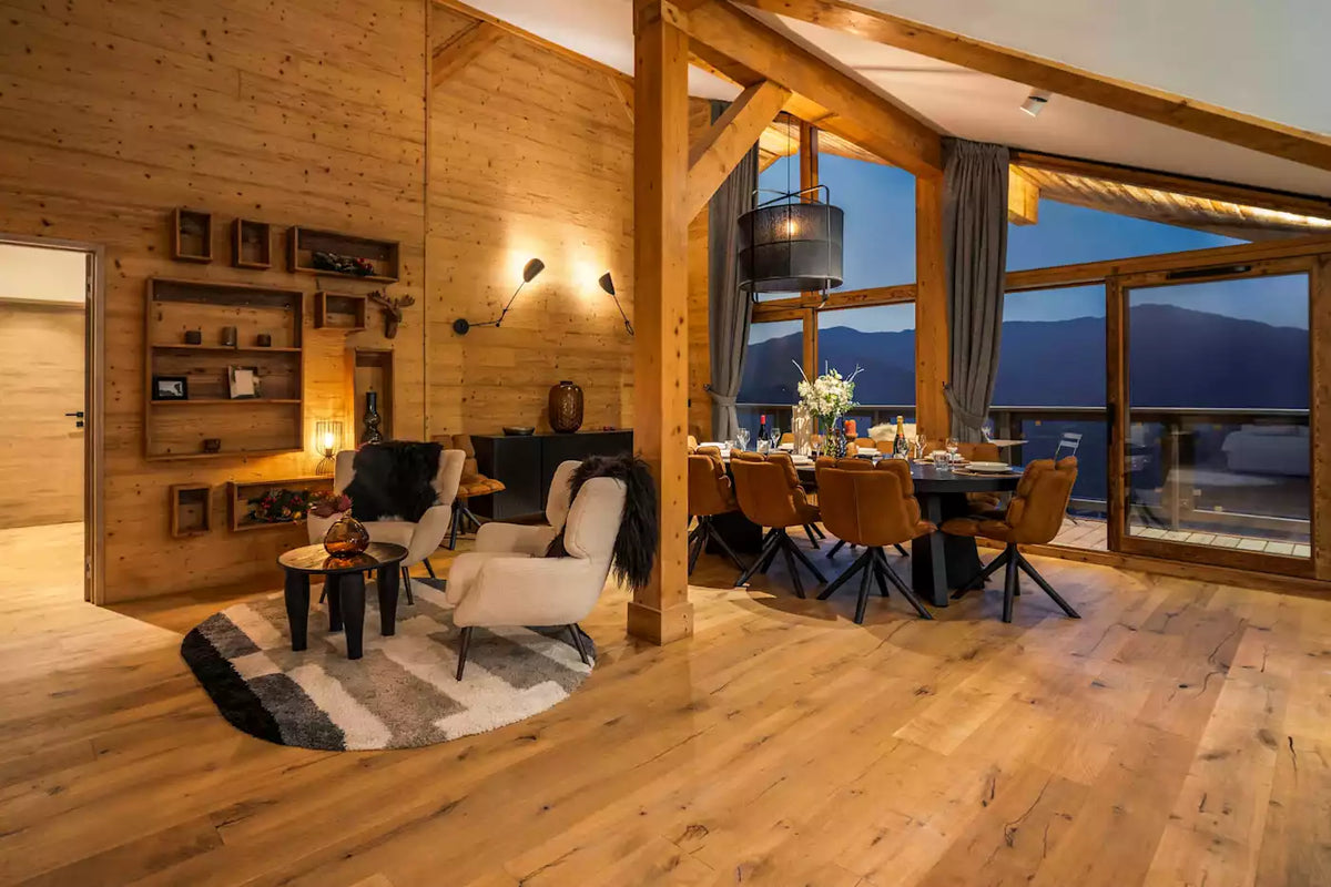 A luxurious mountain chalet interior with warm wood-paneled walls, a dining area with a table and chairs, and a cozy living space with armchairs and a rug, overlooking a mountain view at dusk.