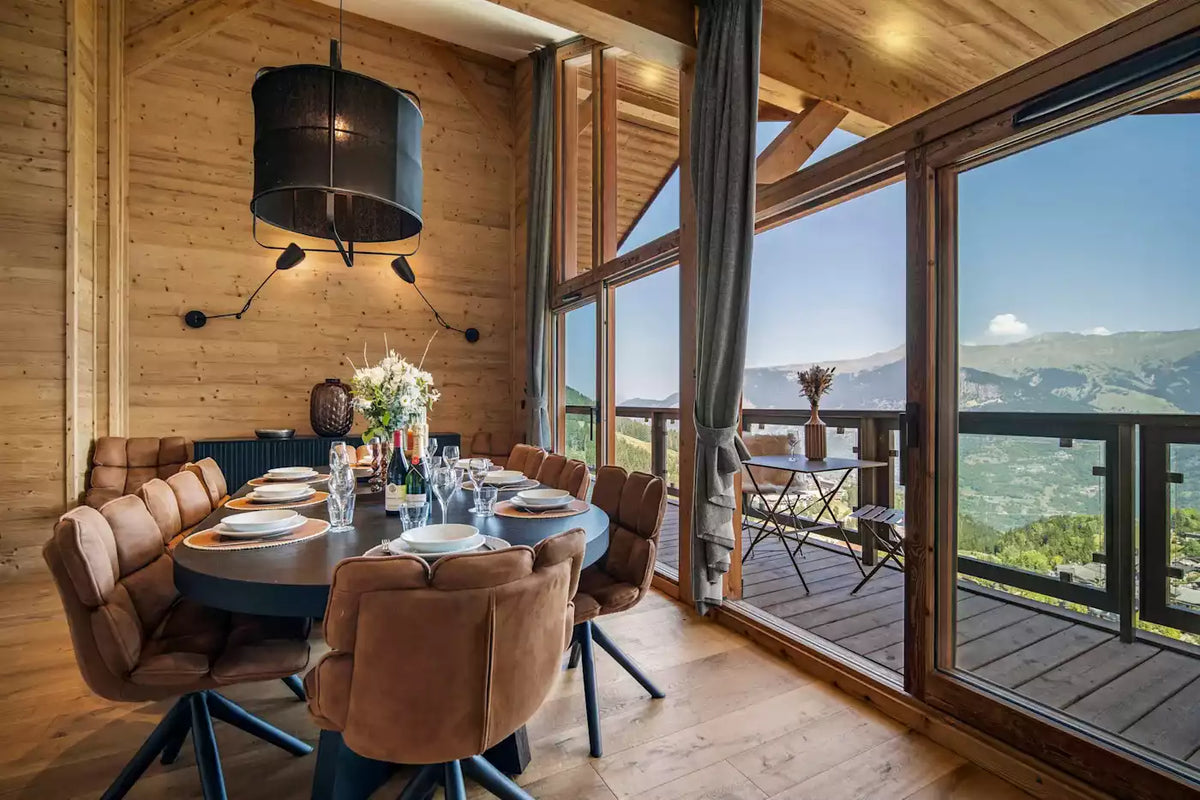 A modern dining room with a round dark table, brown upholstered chairs, and large windows overlooking a mountain landscape. The room has light wood walls and ceiling, a black pendant light, and a small balcony area with a table and chairs.